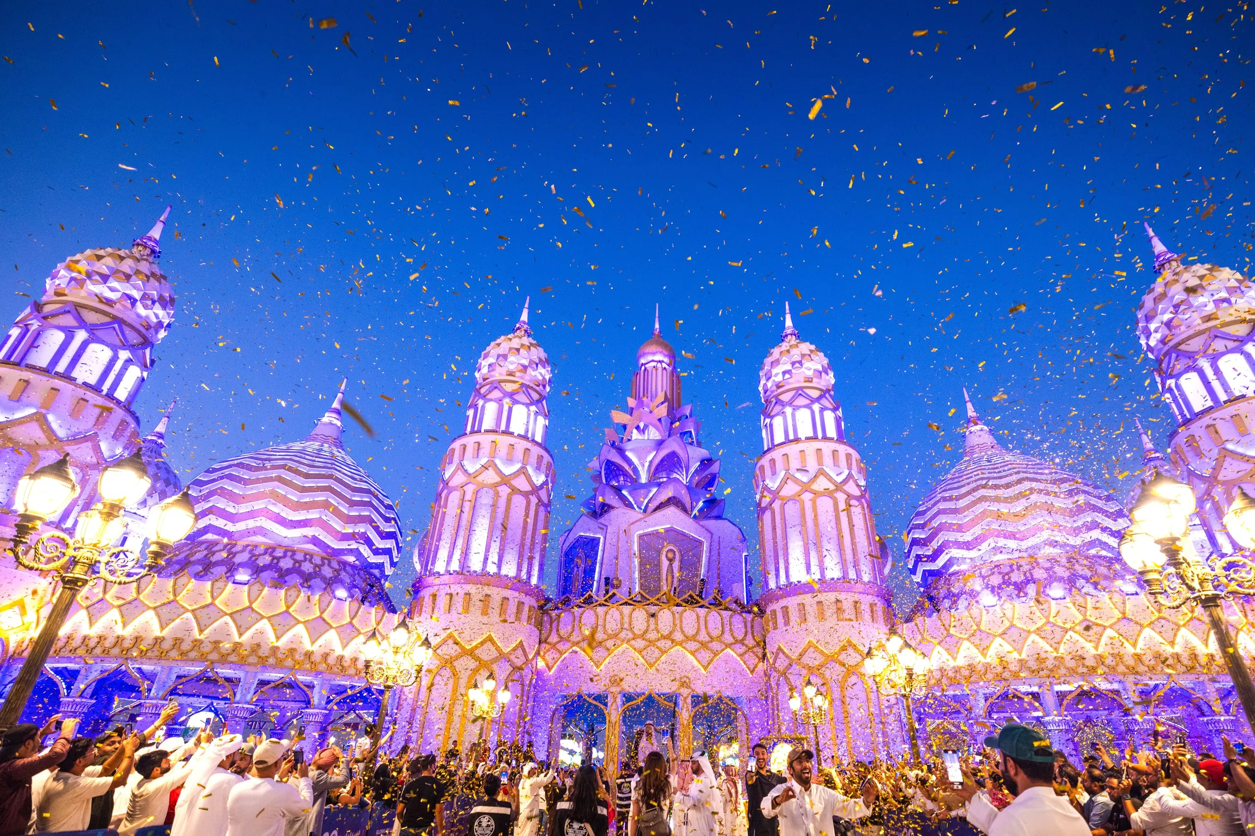 The Global Village gate at dusk under falling gold confetti, with a crowd filling the square in front of it, phones up.