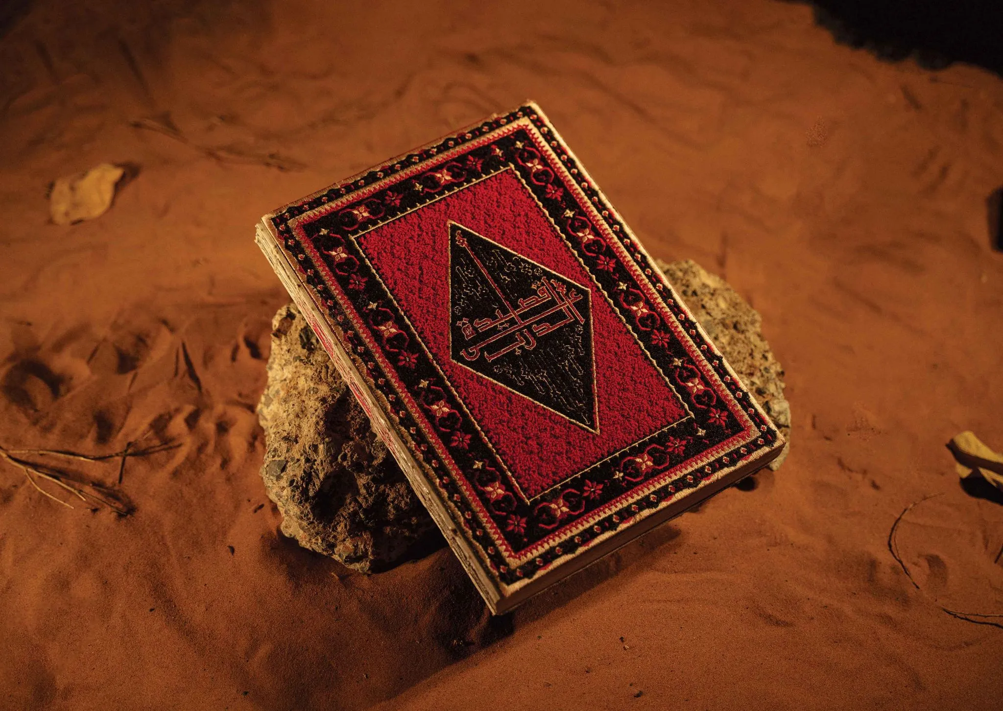 The Poetry In Motion book resting on a rock in desert sand at night, its cover hand-embroidered in red, black and gold with an Arabic title inside a central diamond.